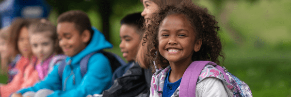 Image depicts elementary age students smiling while wearing backpacks outdoors.