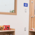 Smiling Black woman childcare worker with short black natural hair, vitiligo, and gray uniform, sitting in an early learning classroom.
